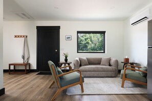 Een moderne woonkamer met een houten vloer, een zwarte deur, een groot raam met uitzicht op bomen, een beige bank en twee groene fauteuils.