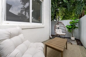 Un sofá blanco acolchado, una mesa de madera, una silla de mimbre y una planta en maceta en una esquina, junto a una valla blanca y follaje.