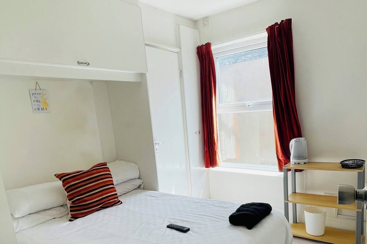 A hotel room with a bed, a bedside table, a red lamp, a window with curtains, and a wall-mounted air purifier.
