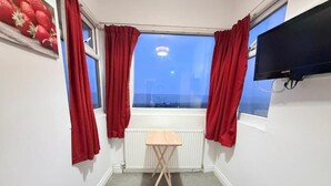 Una stanza con finestra, tende rosse, un piccolo tavolo di legno e un termosifone.