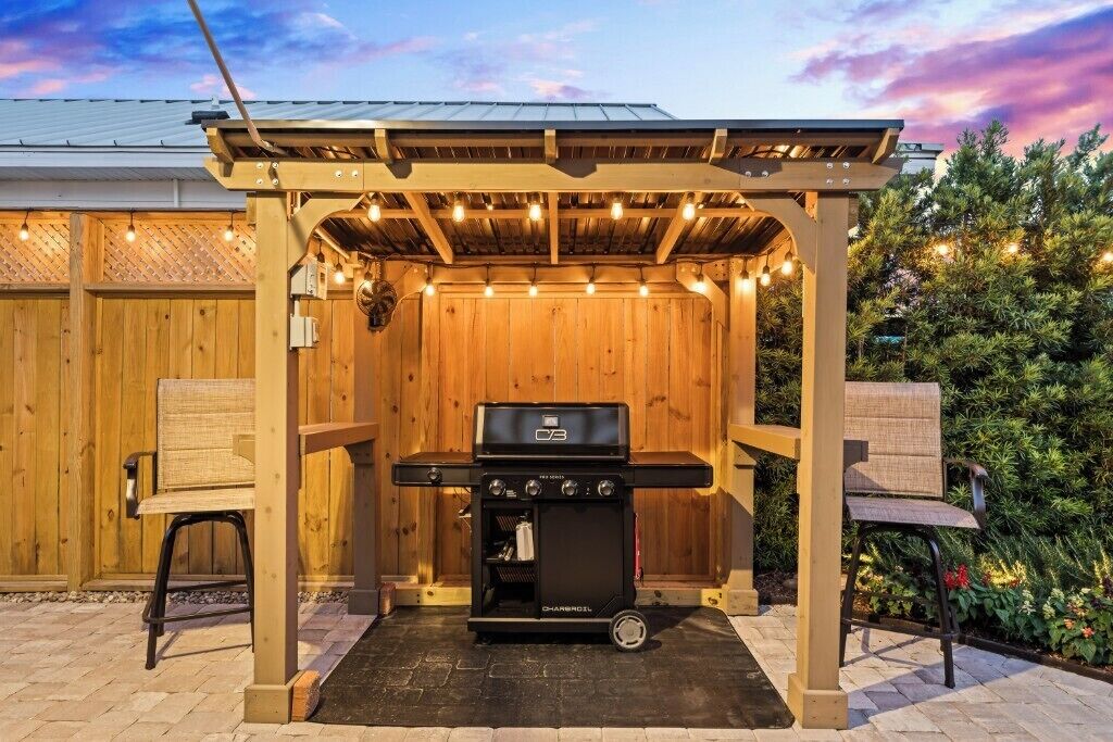 A barbecue grill under a wooden pergola with string lights, surrounded by a wooden fence and a garden.