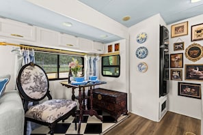 A compact, well-lit interior with a checkered floor, a dining table with blue plates, a decorative chair, and a wall adorned with framed artwork and plates. - French Country Farm Stay RV–Sunsets, Horses & Cozy Cottage Charm in Fairhope, AL (Fairhope)