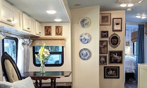 A cozy RV interior with a dining area, a window with a view, and a wall decorated with various framed pictures and plates.