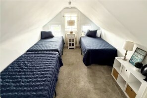 Un pequeño y estrecho dormitorio en el ático con dos camas, una mesita de noche y una lámpara.