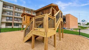 Un parque infantil con estructuras de madera y un edificio con balcones al fondo.