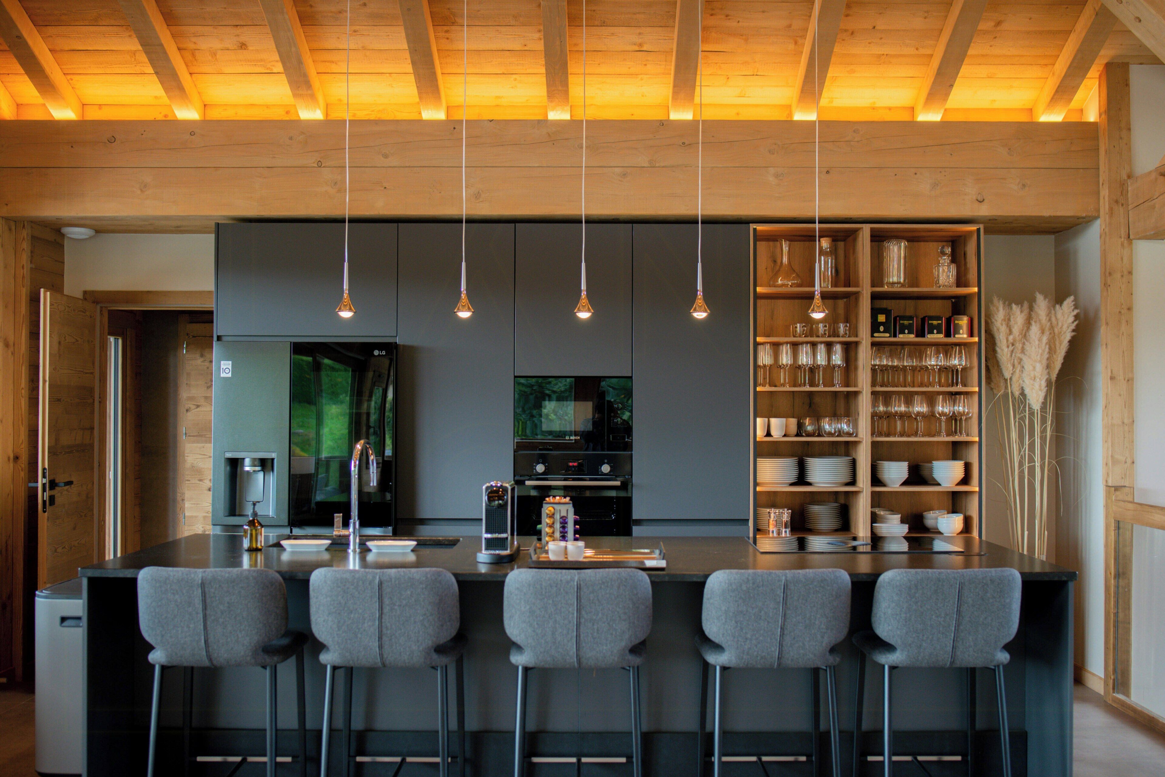 A modern kitchen with a central island, bar stools, hanging pendant lights, and a glass display cabinet.