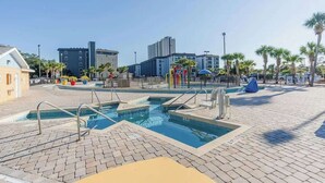 A swimming pool area with multiple pools, a children's play structure, and a fenced playground. - Bright Beachfront Studio + Pool (Myrtle Beach)