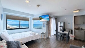 A bedroom with a large window overlooking the ocean, a bed, a desk, and a kitchenette. - Bright Beachfront Studio + Pool (Myrtle Beach)