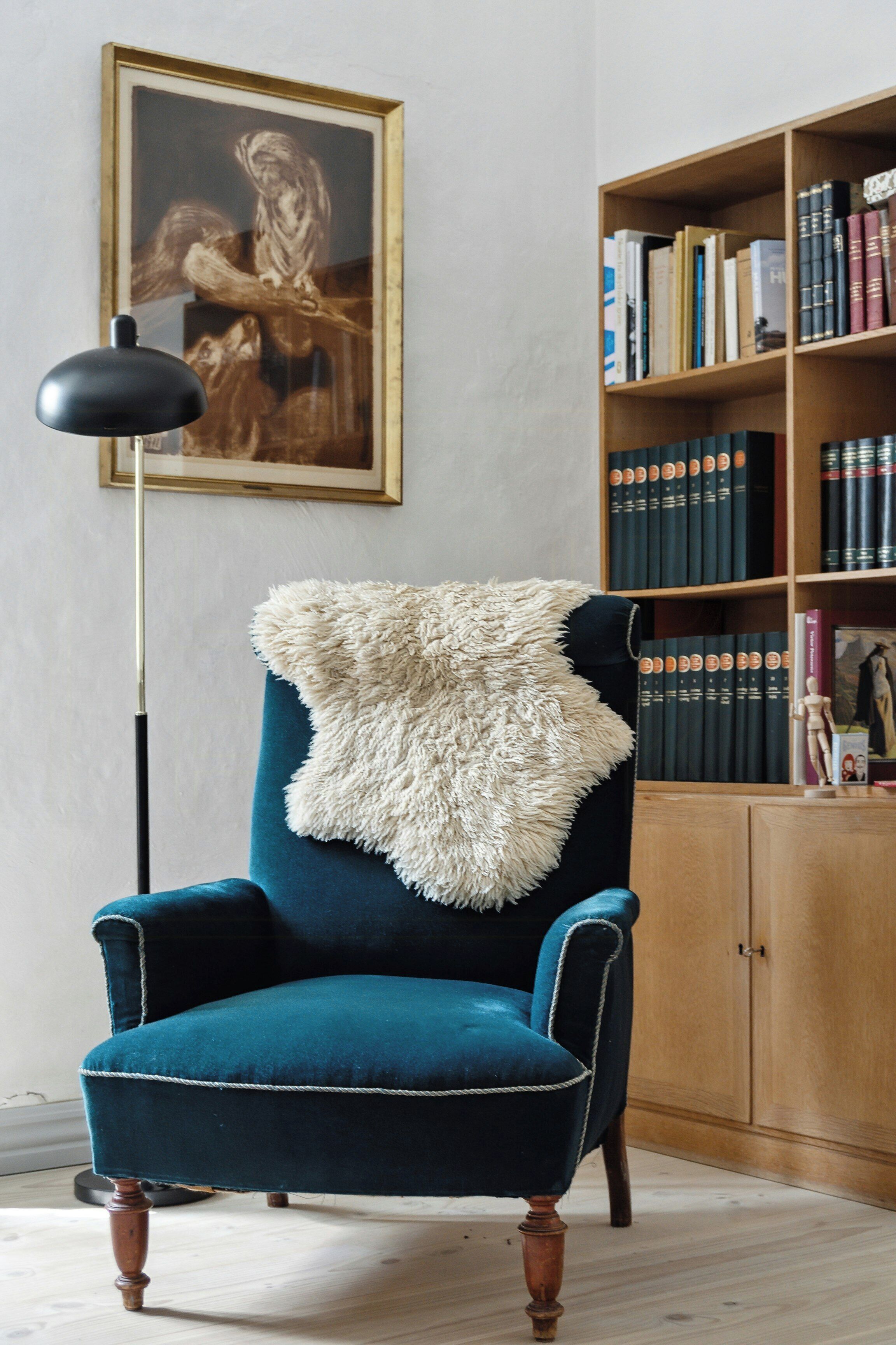 A teal armchair with a fluffy white throw, a floor lamp, and a bookshelf with books and a framed picture.