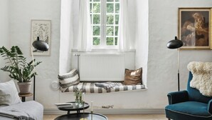 Una habitación acogedora con ventana, radiador, un banco con cojines, una silla, una mesa con vela y una planta.