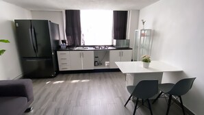 Kompakti keittiö, jossa on jääkaappi, liesi ja pesuallas, ruokapöytä tuoleineen sekä lasiovilla varustettu kaappi.