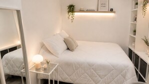 Moderni makuuhuone, jossa on sänky, yöpöytä, lamppu, peili ja hylly, jonka päällä on kasvi ja kehystetty sitaatti.