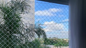 A window with a chain-link fence, a palm tree, and a building in the background. - Oceana Suites Cachoeira Paulista (Cachoeira Paulista)