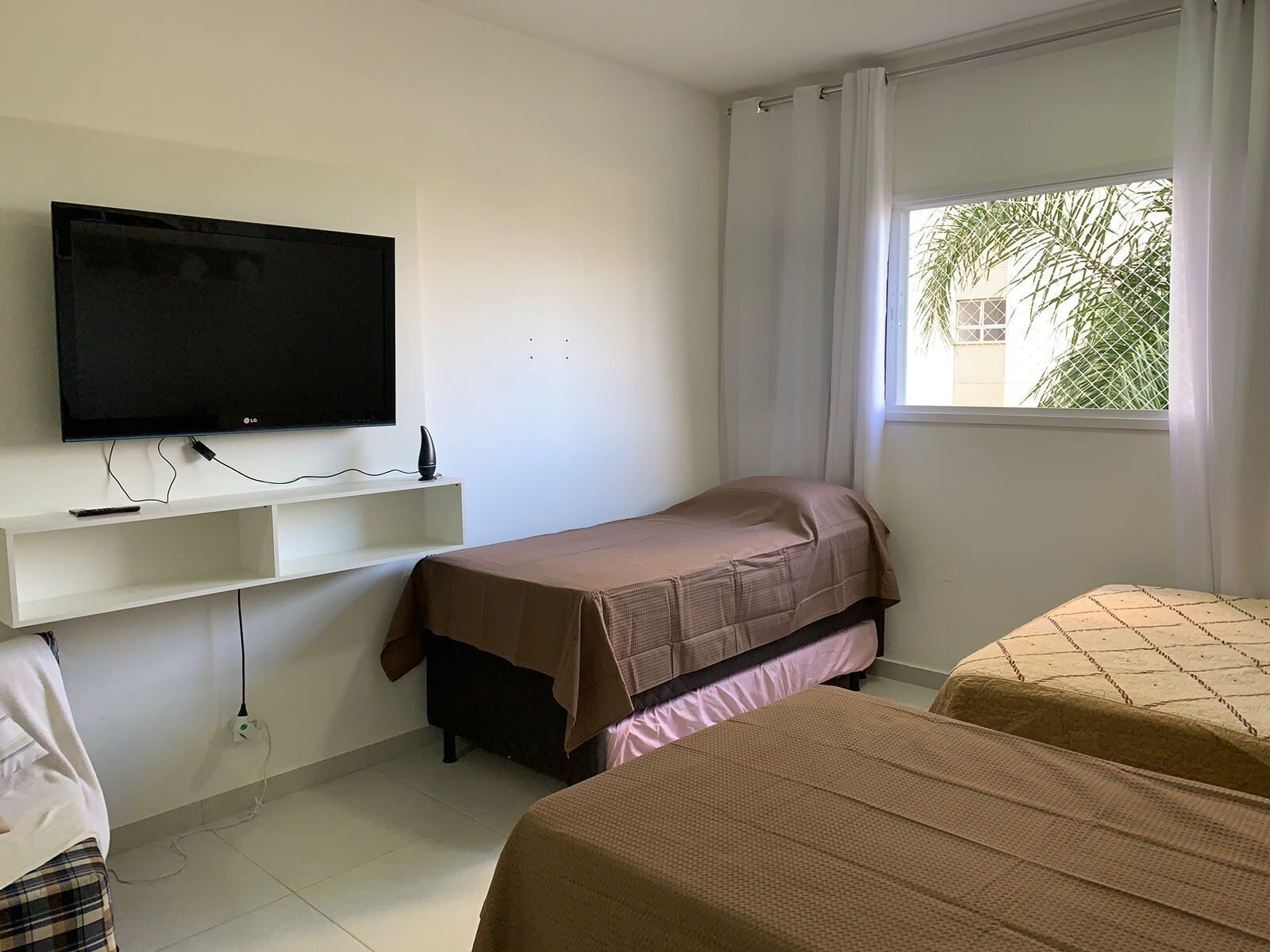 A room with a bed, a TV mounted on the wall, a window with curtains, and a small shelf.