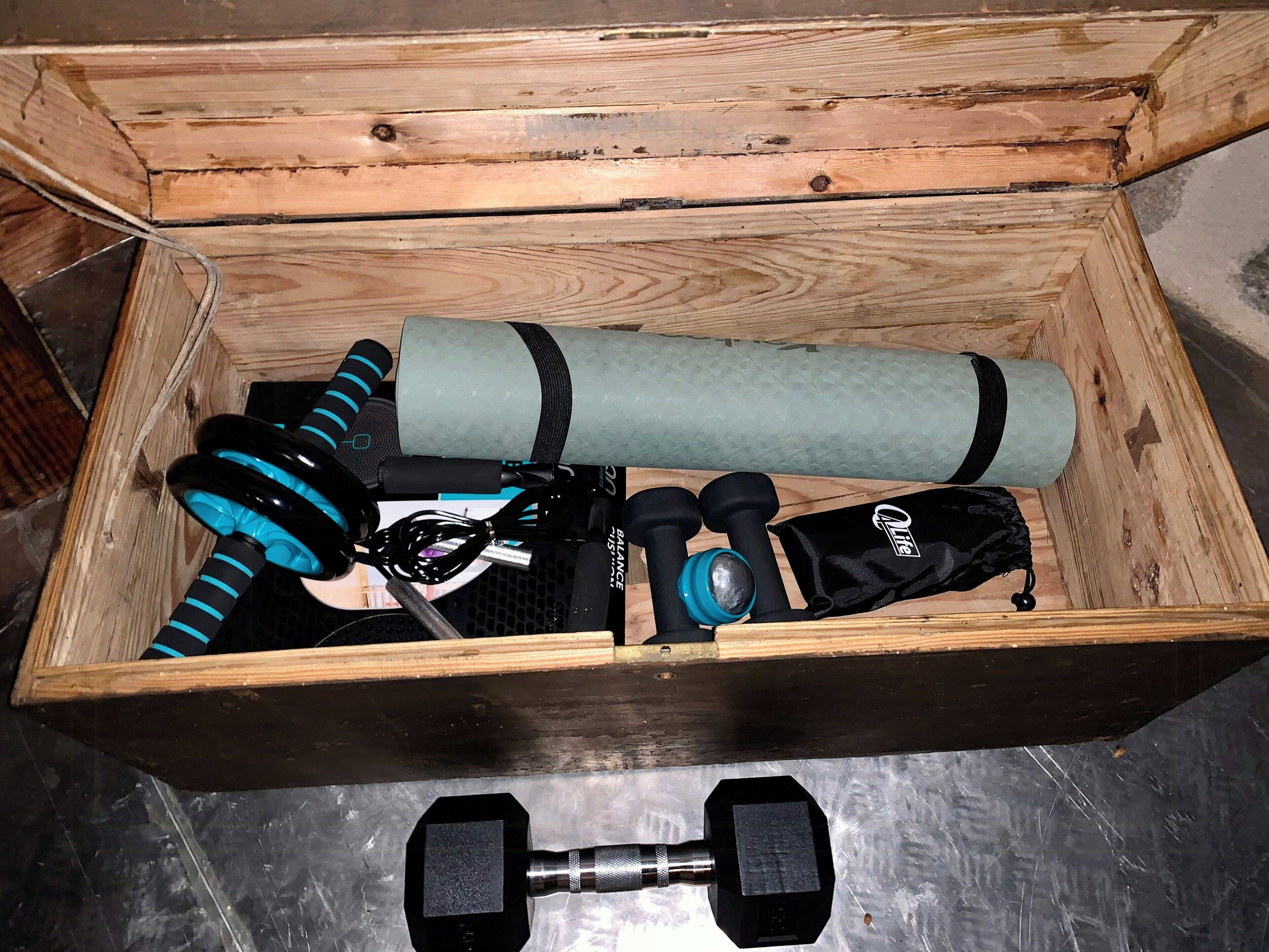 Une boîte en bois contenant du matériel de fitness, notamment des haltères, un tapis de yoga, des bandes de résistance et une bouteille d’eau.
