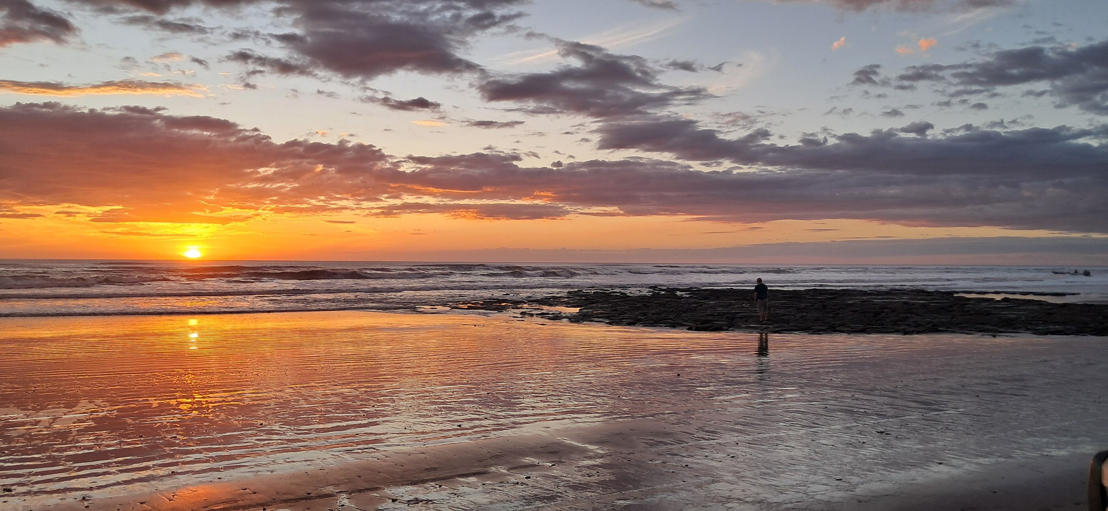 A beach at sunset with waves, a person standing, and a clear sky.