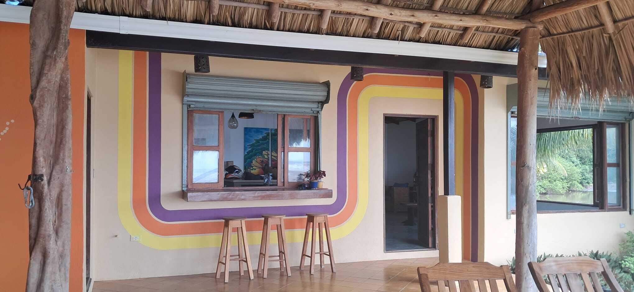 Een kamer met een raam, een houten kruk, een rieten dak en een kleurrijk wandontwerp.