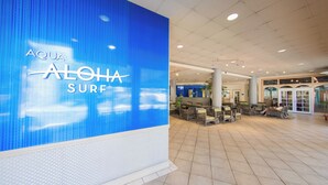 Un hall d’entrée spacieux, avec un grand mur bleu sur lequel est affiché le texte « AQUA ALOHA SURF ». On y trouve des coins salon avec des chaises et des tables, et un comptoir d’accueil en arrière-plan.
