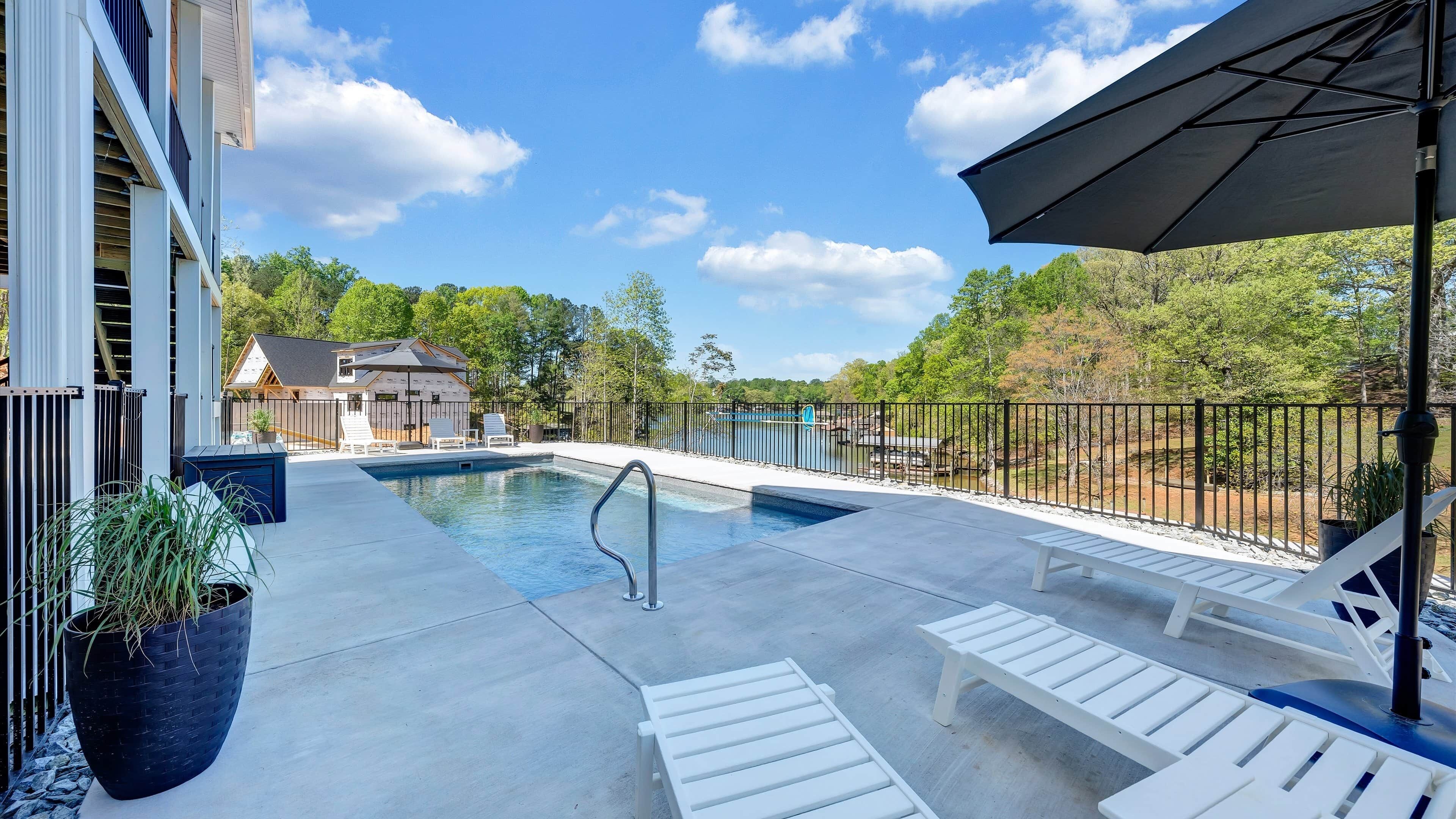 A swimming pool with lounge chairs and a covered area, surrounded by a fence and trees.