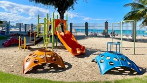 Ein Spielplatz mit Rutschen und Klettergerüsten, umgeben von Sand und einem Zaun.