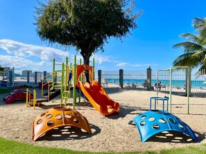Ein Spielplatz mit Rutschen und Klettergerüsten, umgeben von Sand und einem Zaun.
