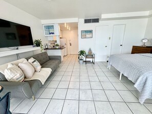 A modern living room with a sofa, a bed, a TV, and a kitchen in the background. - Charming Isla Verde with Pool & Beach Access (Carolina)