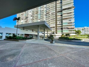 A modern building with a covered entrance, a parking area, and a large multi-story residential structure in the background. - Charming Isla Verde with Pool & Beach Access (Carolina)