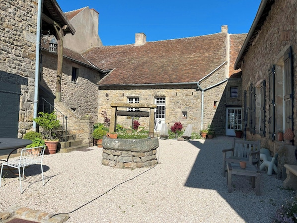 A stone courtyard with a central stone fountain, outdoor seating, and a rustic building with a tiled roof.