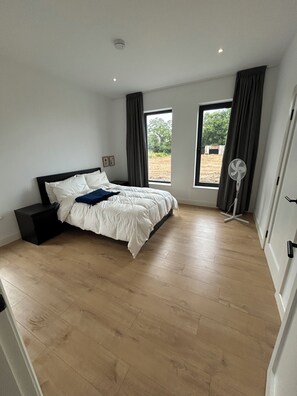 A bedroom with a large bed, a bedside table, a standing fan, and a window with curtains. - Modern Rural Ireland Retreat – Spacious & Peaceful Countryside Escape. (Derryadd)