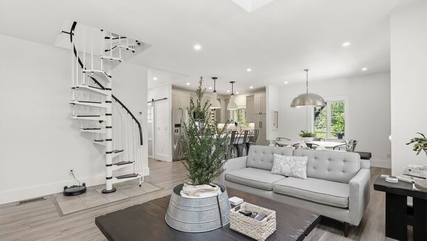 A modern living room with a staircase, a grey sofa, a coffee table, and a dining area in the background. - Designer Home with Wraparound Porches & Fire Pit (Bourne)