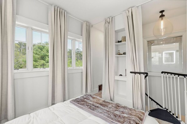 Un dormitorio con cama, ventana con cortinas, radiador, estante con objetos y una escalera.