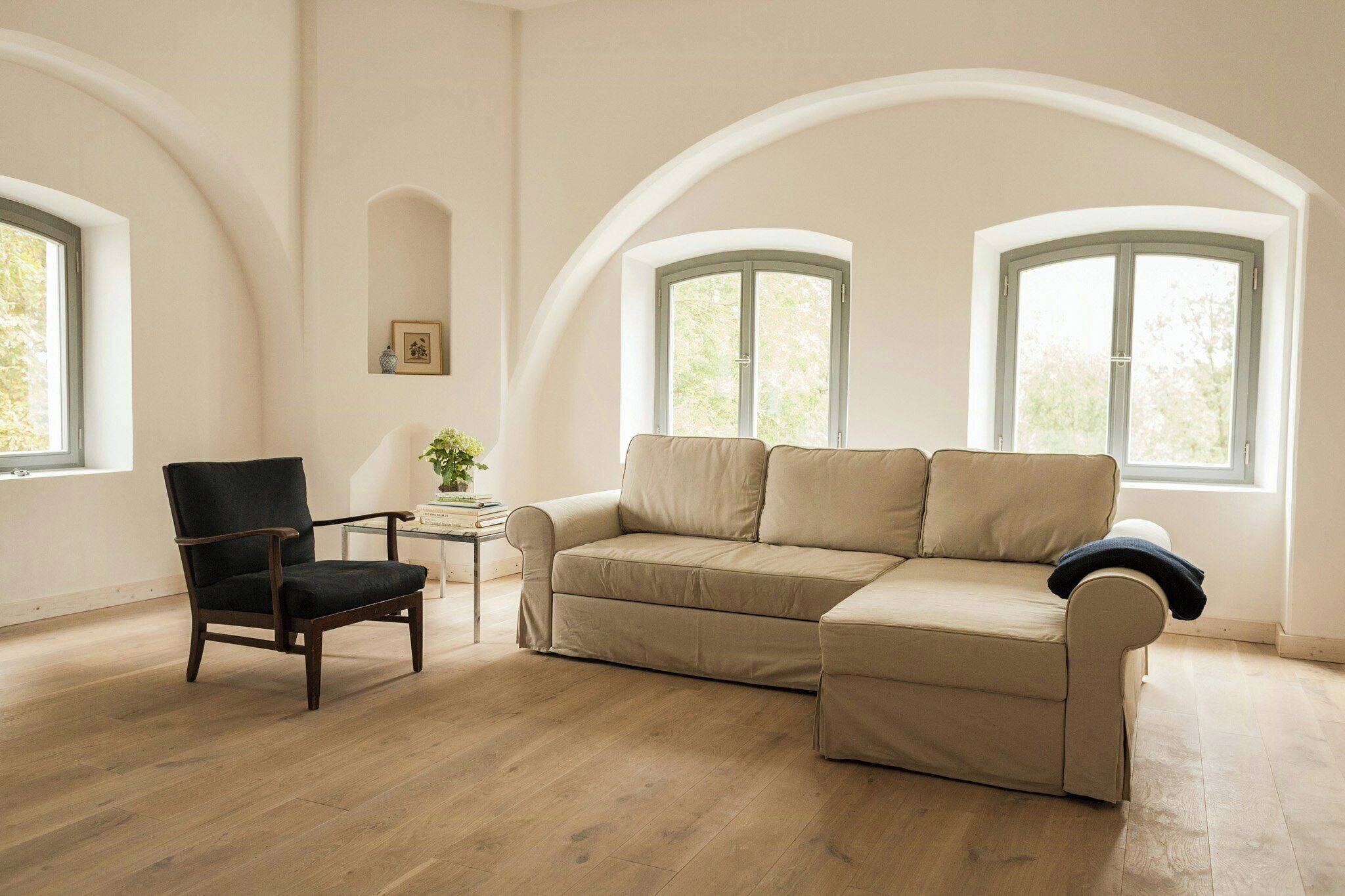 A room with a wooden floor, a black chair, a beige sofa, and two windows with green frames.