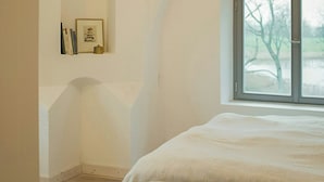 Une chambre avec une grande fenêtre, un lit et une étagère fixée au mur, sur laquelle se trouvent des livres et un tableau encadré.