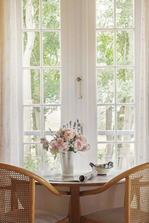 Une pièce dotée d’une grande fenêtre, d’une table en bois, de deux chaises en osier, d’un vase rempli de fleurs et d’un bol posé sur la table.