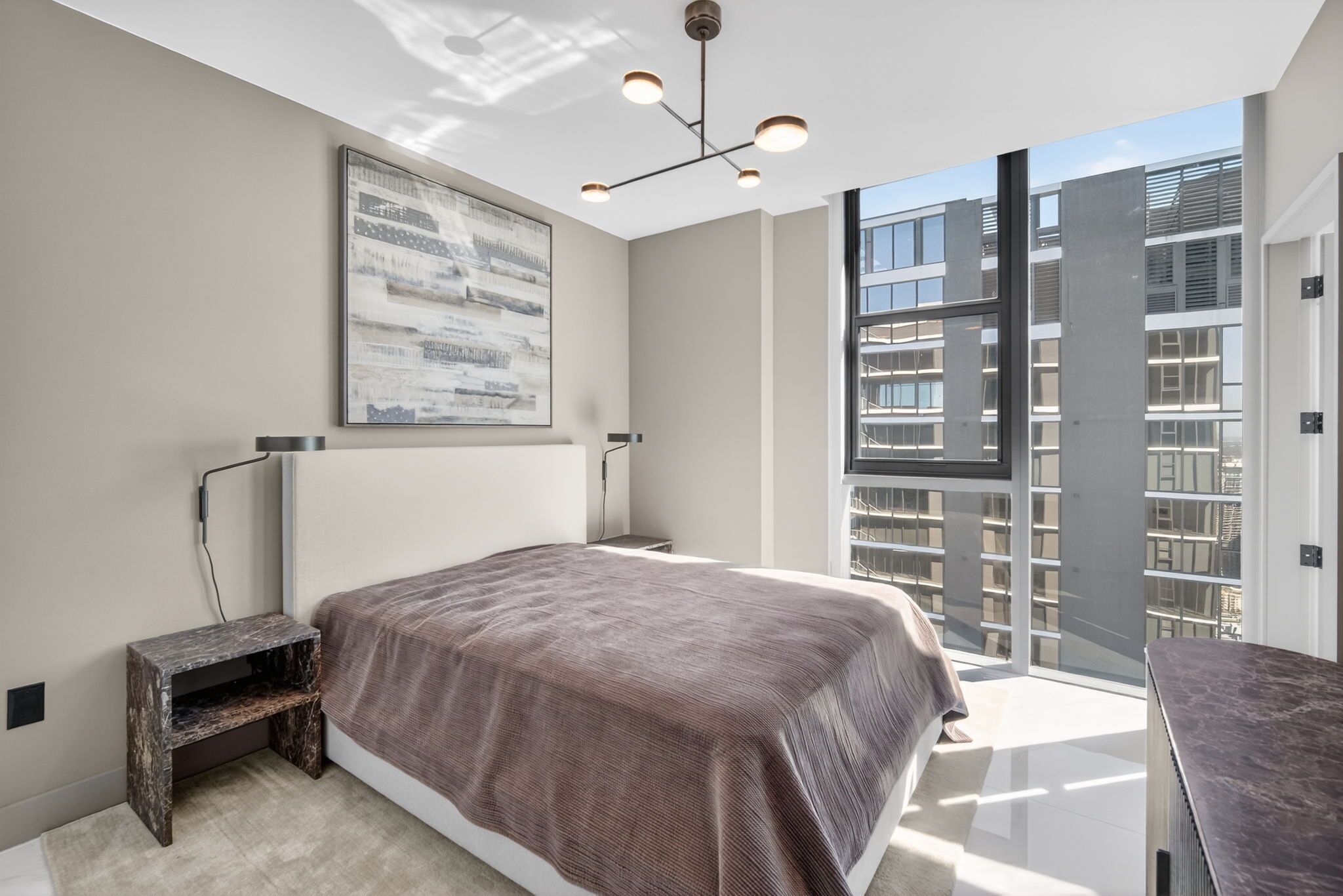 Un dormitorio con una cama grande, una lámpara de araña moderna, un ventanal y vistas a un edificio.