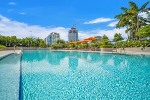 A clear blue pool with lounge chairs and umbrellas, surrounded by greenery and modern buildings. - Waterfront Opulent Sky Penthouse - Aria Reserve (Miami)