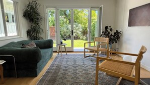 Ein Wohnzimmer mit einem grünen Sofa, einem Holzstuhl, einem gemusterten Teppich und Blick durch Glastüren in einen Garten.