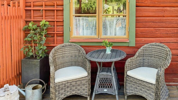 Una cabaña de madera con dos sillas de mimbre y una mesita, una ventana con cortinas y una planta en maceta.