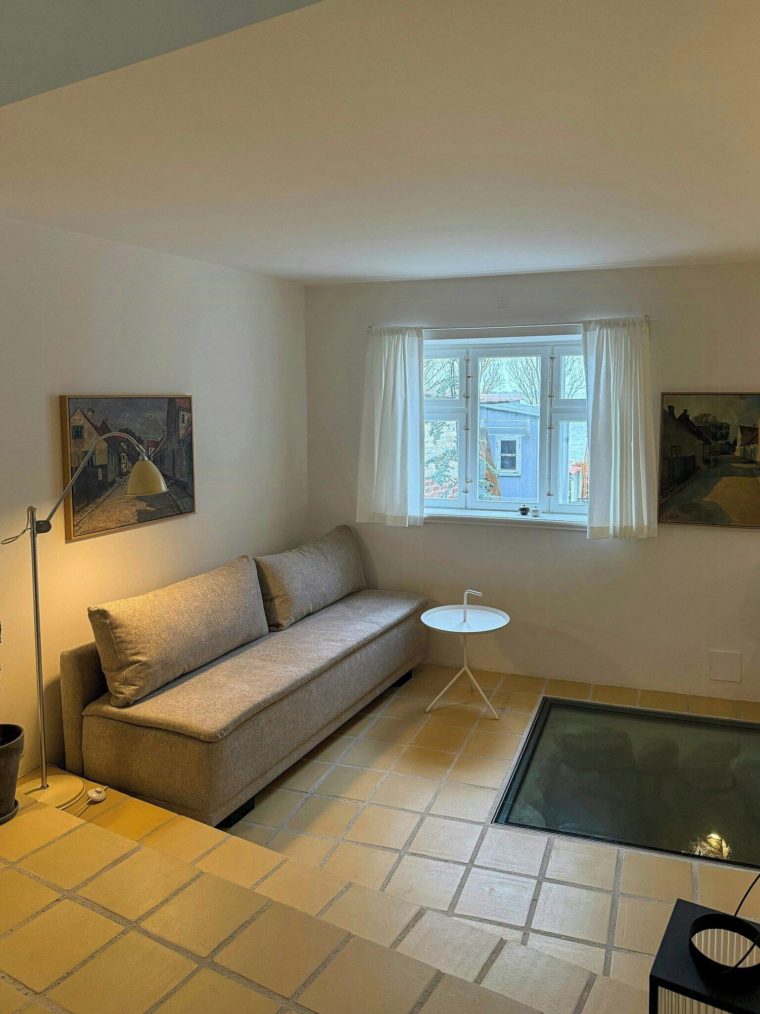 Una sala de estar con un sofá, una mesita redonda, una lámpara de pie y una ventana con cortinas.