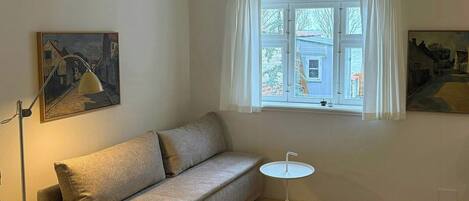 Una sala de estar con un sofá, una mesita redonda, una lámpara de pie y una ventana con cortinas.