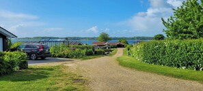 Ein Kiesweg führt zu einem Haus, nebenan ist ein Auto geparkt, umgeben von Grün und mit Blick auf einen See.