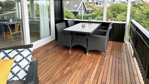 En täckt uteplats med ett bord och stolar, en glasdörr som leder in till ett inomhusutrymme och en utsikt över hus i bakgrunden.