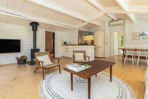 A cozy living room with a fireplace, a dining area with a table and chairs, and a kitchen in the background. - Log cabin with pool near Tisvilde (Vejby)