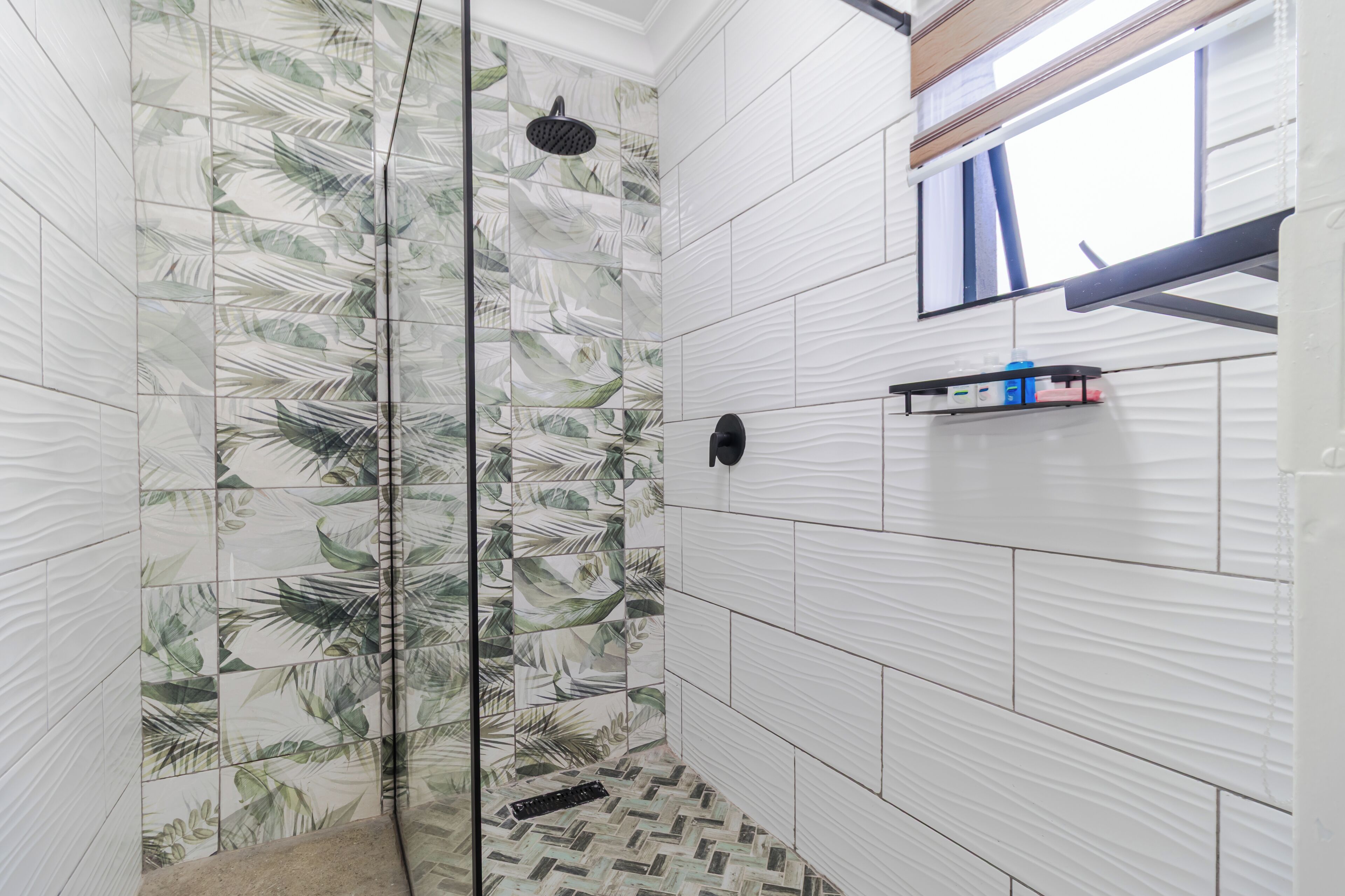 A shower with a glass door, white tiles with a leaf pattern, a small shelf with toiletries, and a window with blinds.