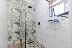 A shower with a glass door, white tiles with a leaf pattern, a small shelf with toiletries, and a window with blinds. - Boutique Hotel-Style Room In Ferndale-H8 (Randburg)