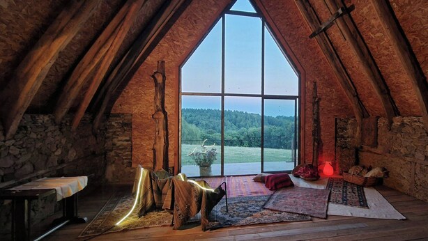 A rustic attic room with a large window, wooden beams, stone walls, and a view of a forest. - The Grange in Cantal (Parlan)