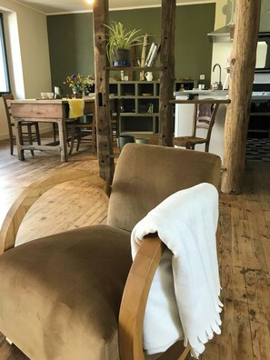 A room with a wooden floor, a brown armchair with a white throw, a wooden table with chairs, a green bookshelf, and a kitchen area in the background. - The Grange in Cantal (Parlan)