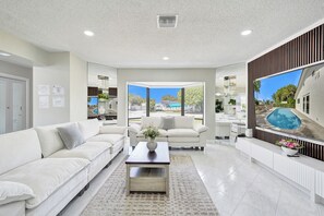 Ein modernes Wohnzimmer mit einem weißen Sofa, einem Couchtisch und einem großen Fernseher, der einen Blick auf einen Pool zeigt.
