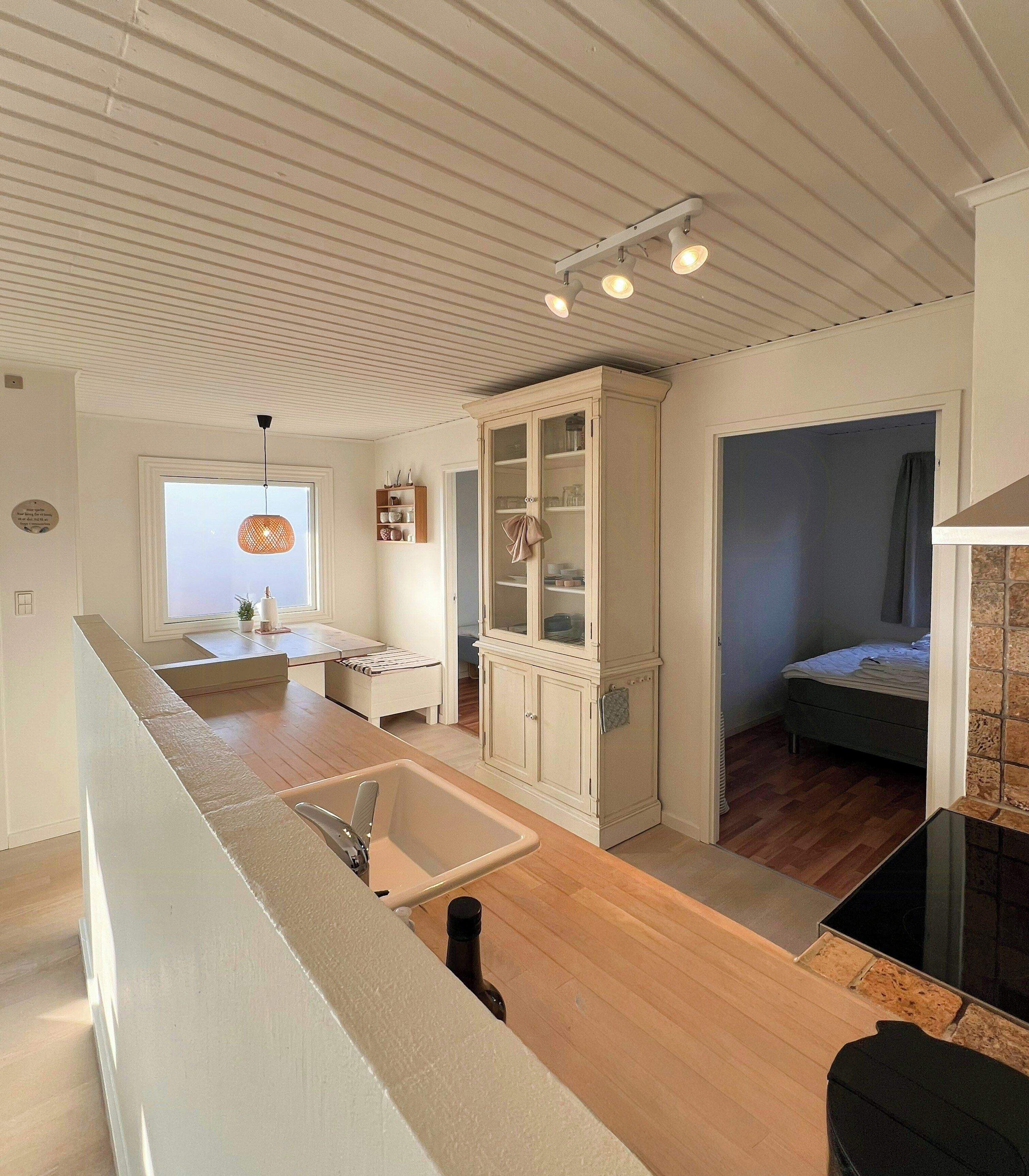 A modern kitchen with a central island, a dining area with a table and chairs, a built-in cabinet with glass doors, and a bedroom with a bed and a window.