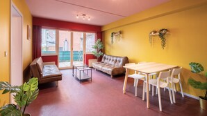Una habitación con sofá, una mesa con sillas, un ventanal de marco rojo y pared amarilla.